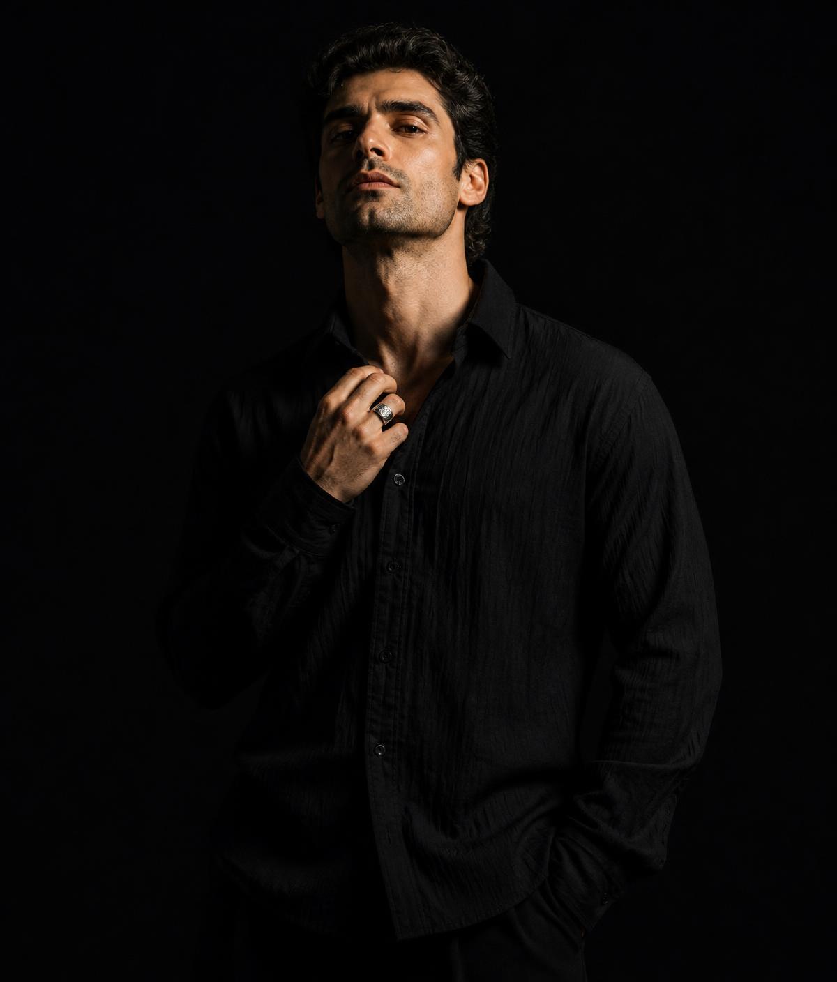 Man adjusting the collar of a black crinkle shirt, silver signet ring visible